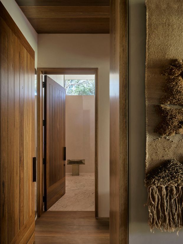 A warm, narrow interior hallway or entry passage featuring extensive dark timber cabinetry and door joinery, a timber-lined ceiling, pale plaster walls, and timber flooring. An open timber door leads to a light-toned bathroom with a stone or marble floor and a small pedestal basin. A textured woven wall hanging adds a natural decorative element on the right.