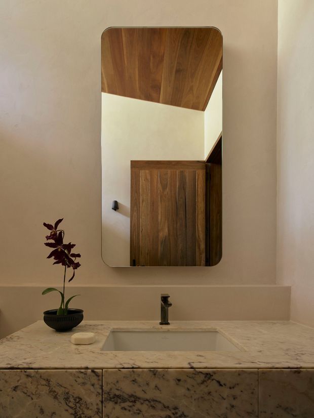 A refined bathroom vanity with a wall-mounted rounded-rectangle mirror above a stone countertop and integrated rectangular basin. The vanity features dark walnut-toned timber cabinetry, while the mirror reflects a warm timber ceiling and adjacent joinery. Soft beige walls, veined natural stone, and subdued lighting create a warm, contemporary atmosphere.