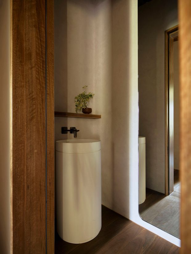 A compact, warmly lit bathroom vanity area viewed through a timber doorway. A freestanding cylindrical white basin sits against a softly textured neutral wall, with a black wall-mounted tap and a narrow timber shelf holding a small plant. Dark timber flooring, timber door surfaces, and a large full-height mirror create a refined, intimate interior.