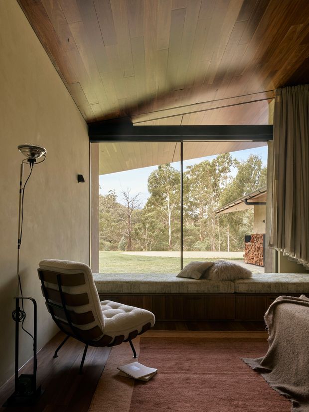 A warm, intimate mid-century-inspired room with a built-in window seat overlooking a leafy garden. The space features a richly timber-lined sloping ceiling, timber flooring and built-in bench joinery, a sculptural upholstered lounge chair, floor lamp, soft curtains and a muted rose-brown rug. Large black-framed glazing and low, cosy proportions create a strong connection to the outdoors.