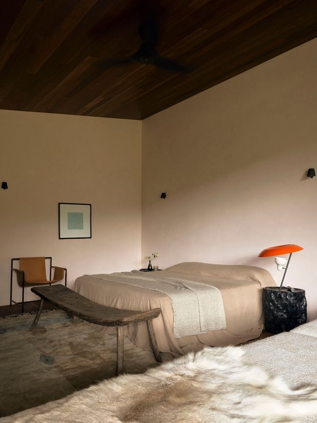 Warm, moody bedroom interior with a low upholstered bed dressed in beige bedding, a textured throw, a rustic dark wood bench, a leather sling chair, and an orange table lamp. The room features a dark timber-lined ceiling with a ceiling fan, softly coloured plaster walls, minimal wall sconces, layered natural-fibre rugs, and restrained contemporary furnishings.