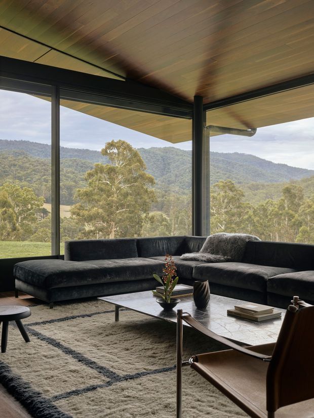 A sophisticated living room with a large dark sectional sofa arranged around a low rectangular coffee table and textured area rug. Floor-to-ceiling black-framed glazing wraps the room, opening views toward wooded hills and a rural landscape. The ceiling is clad in warm-toned timber boards, with a dark structural column and a visible roof gutter at the glazed corner. A leather sling chair, decorative vessels, books, and a small floral arrangement complete the restrained, nature-connected interior.