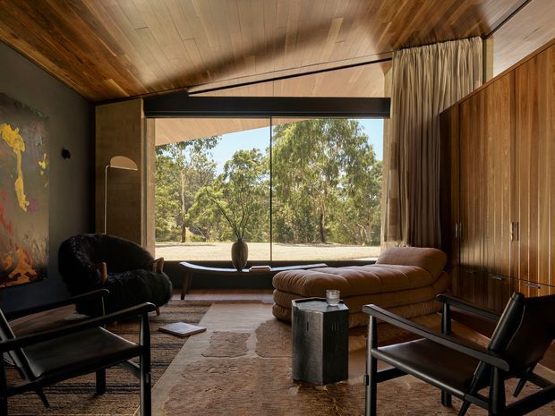 A warm, mid-century-inspired living room with a timber-lined ceiling, dark timber wall panelling and full-height glazing overlooking a wooded landscape. The room contains a low modular brown sofa, black leather lounge chairs, a sculptural floor lamp, textured rug and a small dark side table. A curtain divides the glazed opening, while the rich timber and restrained furnishings create a calm, intimate atmosphere.
