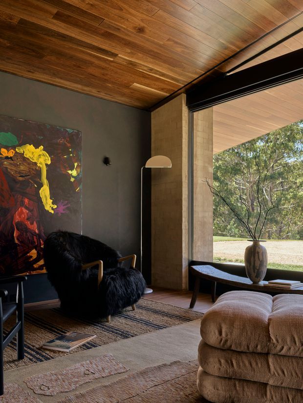 A moody, refined living room with a dark painted wall, richly grained timber ceiling, and large open glazing overlooking trees. The room features a black shag-upholstered lounge chair with timber arms, a slim arched floor lamp, low dark timber coffee table, sculptural ceramic vase, layered neutral rugs, and a large textured beige ottoman. A bold abstract artwork adds a colourful accent. The interior combines warm natural materials with strong indoor-outdoor connection and dramatic contrast.