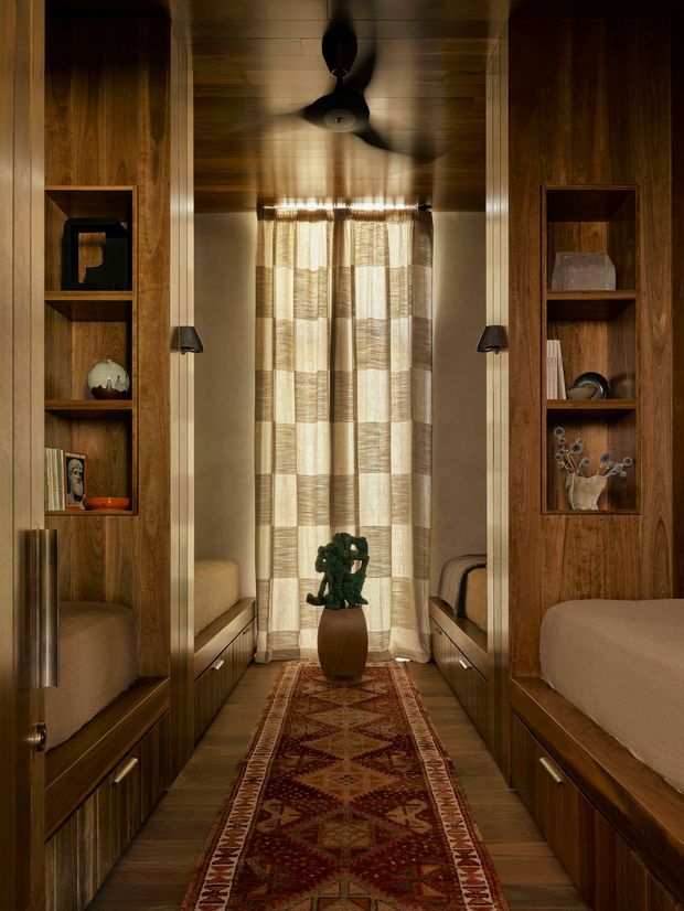 A narrow, warm-toned bedroom or sleeping alcove lined with dark timber built-in cabinetry and open display niches. Two upholstered beds or daybeds flank a central passage leading to a curtained window, with integrated bedside drawers, wall-mounted reading lamps, a ceiling fan, and a patterned runner rug. The space has a richly layered, intimate and highly crafted appearance.