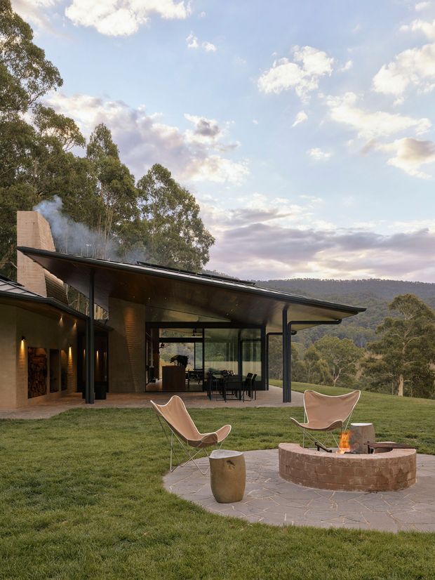 A contemporary single-storey pavilion-style home set in a green, wooded hillside landscape. The building features broad low-pitched roof planes with deep eaves, dark slender structural posts, extensive floor-to-ceiling glazing, warm exposed brick walls, and an open covered outdoor dining area. In the foreground, a circular brick fire pit with a small fire sits on a paved terrace beside sculptural lounge chairs, with expansive lawn and eucalyptus-covered hills beyond.