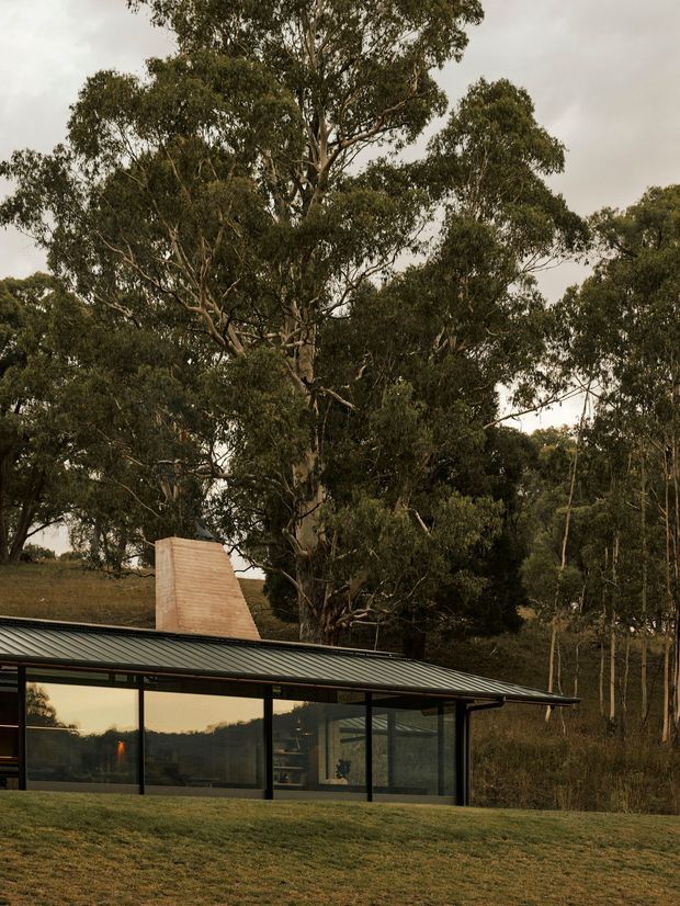 A low-slung modern pavilion set within a wooded rural landscape. The building features extensive floor-to-ceiling glazing framed in dark metal, a shallow dark pitched roof with broad eaves, and a prominent sculptural brick chimney rising behind the glazed living space. Mature eucalyptus trees and grassy terrain surround the structure, creating a strong connection to the bush setting.