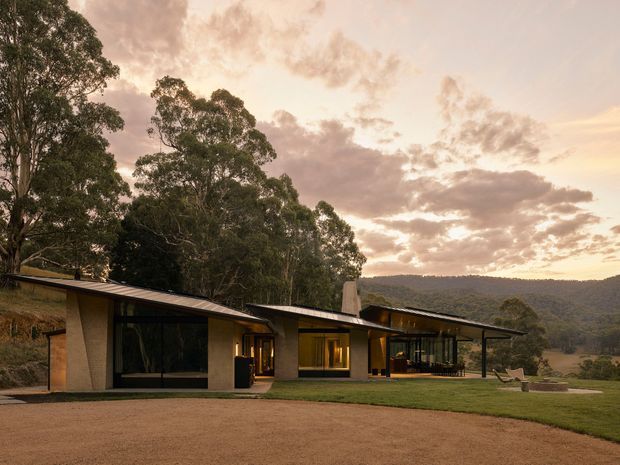 Single-storey contemporary residence set within a wooded, hilly landscape at sunset. The long, low building has broad dark-framed glazing, warm-toned rendered walls, projecting shallow metal roofs, and connected wings arranged around a manicured lawn. A gravel driveway or forecourt occupies the foreground, with outdoor seating visible near the glazed living area.