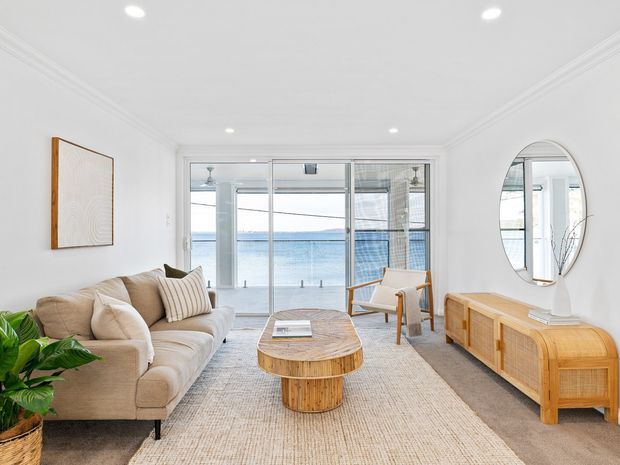 Bright coastal living room with a beige upholstered sofa, woven rug, rounded timber coffee table, rattan-style timber sideboard, armchair, oval wall mirror, and indoor plants. Full-height glazed doors open the room to a balcony with a visible water view. White walls, ornate ceiling cornices, and recessed lighting create an airy, refined interior.