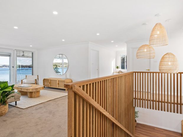 Bright, airy open-plan residential interior viewed from an upper landing, with a timber slatted stair balustrade in the foreground. The living area includes neutral carpet, a curved timber coffee table, upholstered seating, a timber media console, and large glazing overlooking water. Woven pendant lights hang above the stairwell, with white walls, ceiling, and trim throughout.