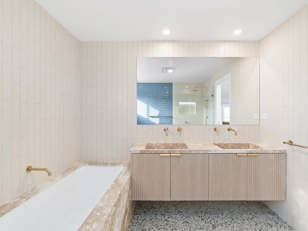 Bright contemporary bathroom featuring full-height white vertical wall tiles, a freestanding-style built-in bathtub with stone-look surround, a double vanity with pale timber cabinetry and a mottled stone benchtop, brass tapware and towel rail, a large mirrored wall cabinet, and terrazzo-style flooring. A blue-tiled adjoining wet area is visible in the mirror reflection.