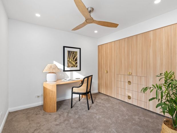 A bright, contemporary bedroom with a compact built-in study desk, woven chair, bedside-style lamp, framed botanical artwork, soft grey carpet, and a full-height light timber built-in wardrobe with integrated drawers. A ceiling fan and potted plant add natural accents.