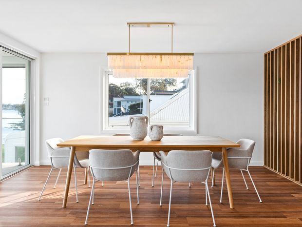 Bright contemporary dining area with a long light-wood dining table surrounded by upholstered chairs. The room features dark timber flooring, white walls, a linear timber pendant light, and a vertical timber-slat screen on the right. Large windows and glazing provide abundant natural light and views outside.