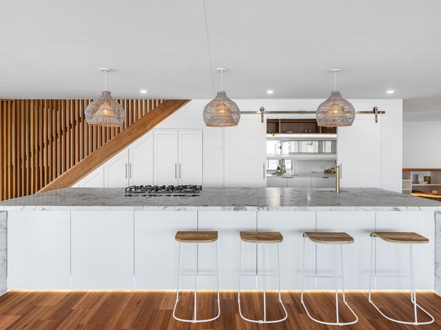 Bright contemporary kitchen featuring a large white marble-look island with four timber-topped bar stools, white integrated cabinetry, a gas cooktop, open shelving, and a striking curved timber slatted staircase screen. Woven pendant lights hang above the island, while warm timber flooring adds contrast.