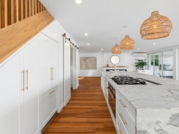 Bright, spacious contemporary kitchen with white shaker-style cabinetry, long marble-look island, integrated gas cooktop, warm timber flooring, and woven rattan pendant lights. A timber stair and balustrade feature is visible at the left, with an adjoining open living area beyond.