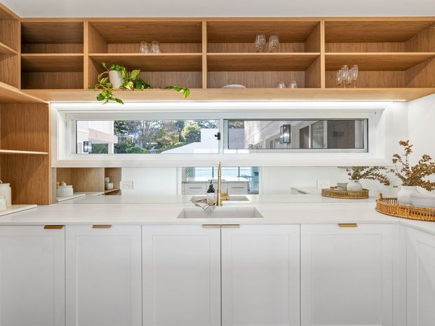 Bright contemporary kitchen featuring extensive white lower cabinetry, a pale engineered-stone benchtop, integrated sink and tapware, and warm timber open shelving with concealed under-shelf lighting. A horizontal window provides natural light and views to the exterior, while restrained plants and tableware add subtle styling.
