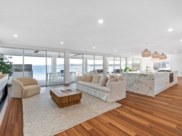 Bright open-plan waterfront living area with a contemporary kitchen and living room. The space features expansive floor-to-ceiling glazing, white cabinetry, a large veined stone island, warm timber flooring, a neutral sofa and armchair, woven pendant lights, and broad water views beyond the glass doors.