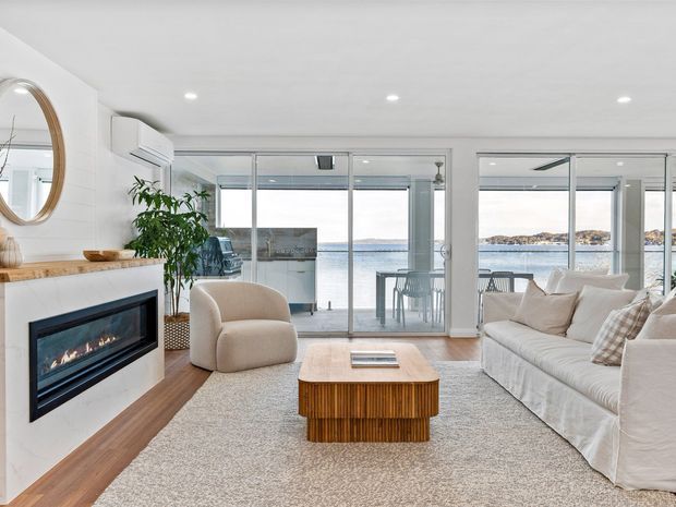 Bright contemporary living room with a large cream sectional sofa, curved upholstered armchair, fluted timber coffee table, and a built-in linear fireplace set into a white feature wall. Full-height glazed doors open to a balcony or terrace with expansive waterfront views. The room has recessed ceiling lights, light carpeting, warm timber flooring accents, and a neutral coastal palette.