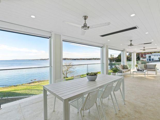 Bright waterfront-facing covered outdoor living and dining area with expansive open glazing, white ceiling, ceiling fans, a slatted dining table with chairs, and a separate lounge setting. The space overlooks calm water and a landscaped shoreline, creating an airy contemporary coastal setting.