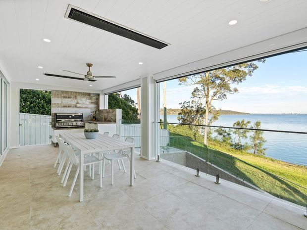 Bright enclosed outdoor entertaining area overlooking a calm waterfront. The space features a long white dining table with multiple white chairs, a built-in stainless-steel barbecue against a textured timber-look feature wall, pale tiled flooring, white-lined ceiling with recessed lighting and ceiling fans, and extensive frameless glass balustrading and glazing. A landscaped waterfront setting is visible beyond the open sides.