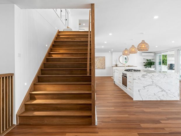 Bright open-plan interior featuring a wide timber staircase with matching timber flooring, leading beside a white contemporary kitchen. The kitchen includes a large white marble-look island, white cabinetry, pendant lights with woven shades, recessed ceiling lighting, and generous glazing.