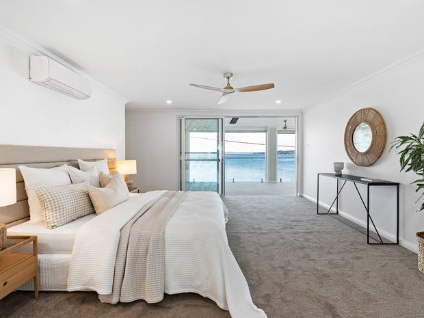Bright, spacious bedroom with a large upholstered bed, layered white and beige bedding, bedside tables, carpeted floor, and minimalist console styling. Wide glazed doors open toward a balcony or terrace with a visible waterfront beyond. White walls and ceiling feature simple cornice detailing, recessed downlights, an air-conditioning unit, and a ceiling fan.