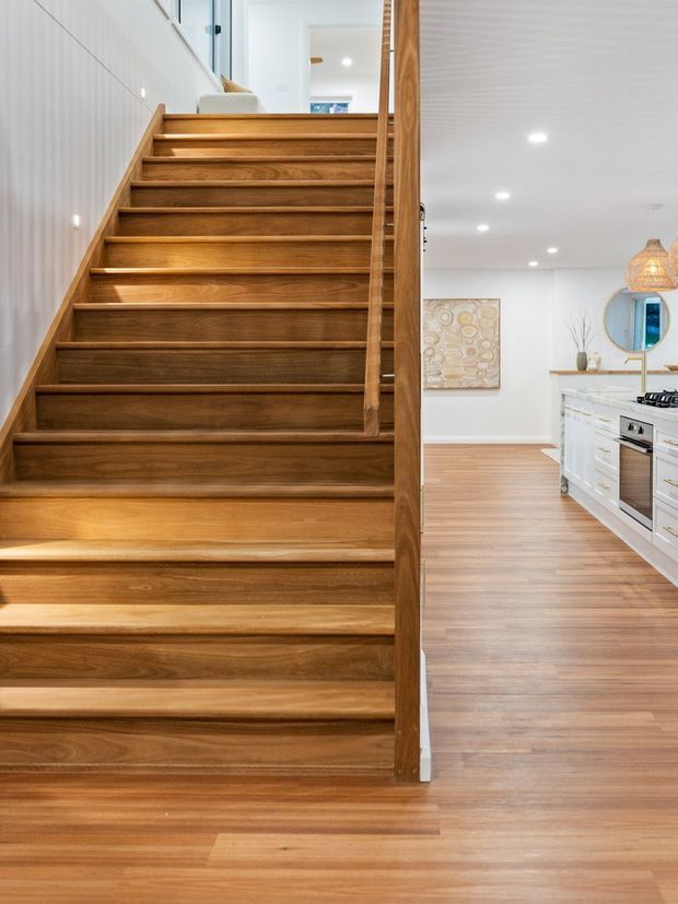 Bright residential interior showing a feature timber staircase with open risers and a timber balustrade, leading to an upper level. A white kitchen with island, cooktop and cabinetry is visible to the right, with matching timber flooring throughout. The space has white walls, recessed ceiling lights, and warm natural wood finishes.