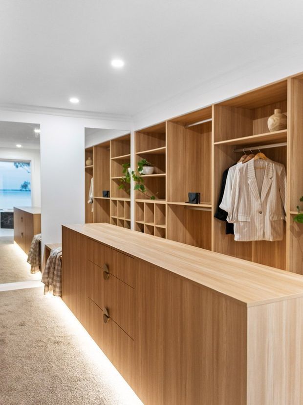A bright, contemporary walk-in wardrobe with extensive custom timber joinery, open shelving, hanging rails, drawers, and integrated lighting. A central island provides additional storage, while a full-height mirror and glimpses of an adjoining bedroom are visible at the left.