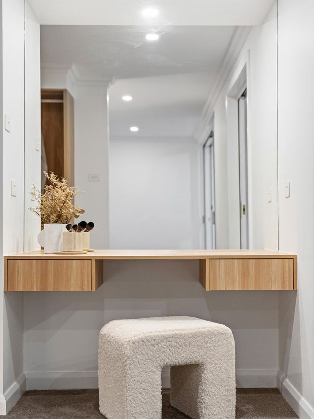 Bright, minimalist hallway dressing area with a large wall-mounted mirror, floating light-wood vanity console, and a plush off-white boucle stool. White walls, recessed ceiling lights, and a carpeted floor create a clean, understated interior.