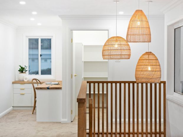 Bright, white-toned interior showing a landing or hallway overlooking a stairwell, with a timber balustrade and three large woven rattan pendant lights. A compact white work or study nook with cabinetry, desk surface, chair and window is visible to the left. The space has pale carpet, white walls and ceiling, and warm timber accents.