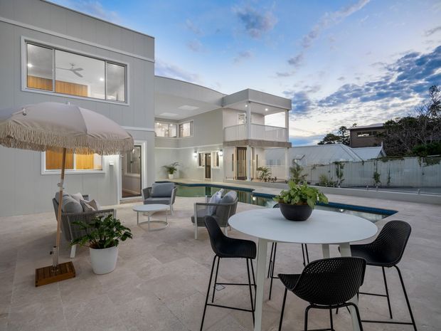 Contemporary multi-level residential courtyard viewed from an outdoor entertaining area, with pale rendered and metal-clad walls, extensive glazing, a covered balcony, a swimming pool, and a round outdoor dining setting beneath a large fringed umbrella. The space has a refined, resort-like feel with neutral finishes and landscaped planting.