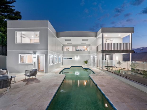 Twilight exterior view of a modern multi-level residence arranged around a long, narrow swimming pool courtyard. The building has crisp white and grey rendered walls, flat roofs, extensive glazing, upper-level balconies and a central open-air courtyard, with outdoor seating and paved pool surrounds.
