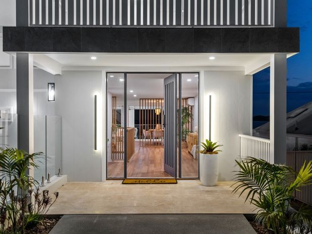 Contemporary residential entry facade photographed at dusk, featuring a covered porch, pale rendered walls, dark charcoal fascia, vertical metal screening, glazed double entry doors, and tropical planting in built-in garden beds. Warm interior lighting reveals timber flooring and a furnished living area beyond the entrance.