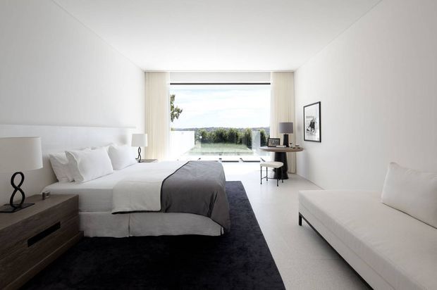 Bright, minimalist bedroom with a low white bed, layered white bedding, charcoal grey throw and rug, a streamlined dark timber bedside unit, and a white upholstered sofa. Floor-to-ceiling glazing at the far end opens to a balcony or terrace with a distant landscape view. A compact desk and chair sit beside the window, with simple monochrome artwork and lighting.
