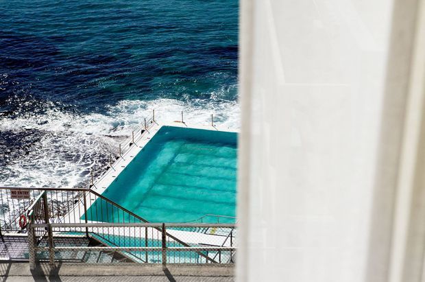 View through sheer white curtains toward a turquoise ocean pool directly beside the surf, with metal safety fencing, pool access stairs and railings visible in the foreground. The image is dominated by the outdoor pool and dramatic coastal setting.