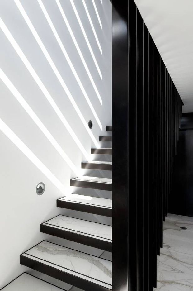 A dramatic contemporary stairway with floating marble-look treads and dark riser or edging details. A tall black vertical slatted screen runs alongside the stairs, contrasting with the white wall and creating strong linear shadows. Recessed circular wall lights illuminate the stairwell, while a pale veined stone floor is visible beyond the screen.