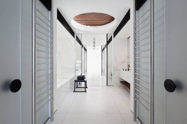 A bright, highly refined interior hallway viewed through open double doors with horizontal metal detailing. The corridor has large pale floor tiles, white marble-like wall surfaces, black-framed glazed partitions, a built-in bench, and a dramatic circular ceiling opening with a timber-lined inset. The space feels minimalist, luxurious, and gallery-like.