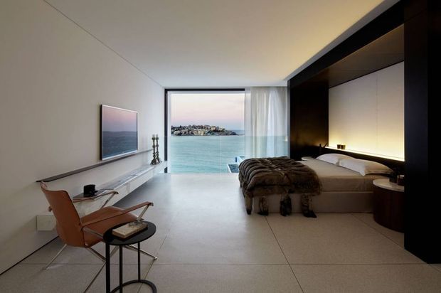 A refined contemporary bedroom with a strong waterfront outlook. The room features a low platform bed integrated into a dark built-in wall, warm concealed bedside lighting, pale large-format tiled flooring, a minimalist desk and leather lounge chair, and floor-to-ceiling glazing opening to a calm blue-water view.