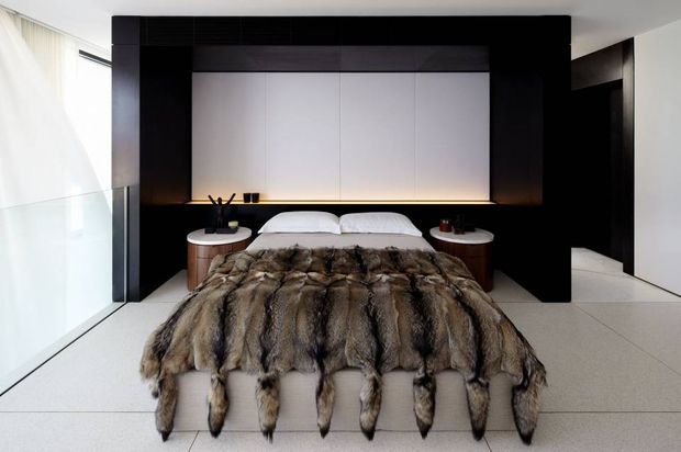 A dramatic contemporary bedroom with a low platform bed framed by a large black built-in feature wall. The headboard incorporates pale vertical panels and warm concealed lighting, with integrated bedside surfaces and minimalist styling. A luxurious fur-like throw covers the bed, while pale flooring, sheer curtains, and dark architectural joinery create strong tonal contrast.