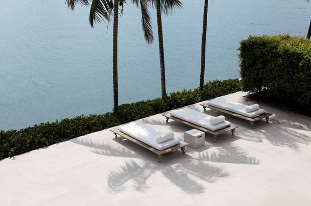 A minimalist waterfront outdoor lounging area with three white cushioned sun loungers arranged on a pale paved terrace beside calm blue water. Tall palm trees, clipped hedges, and strong palm shadows create a refined tropical resort atmosphere.