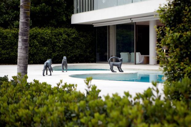 A landscaped modern residential exterior viewed through foreground hedging, featuring a pale paved pool terrace, a turquoise swimming pool, sculptural animal figures, dense greenery, and a contemporary two-storey facade with extensive floor-to-ceiling glazing and white horizontal slab overhangs.