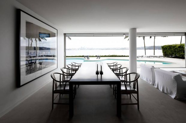 A refined dining room overlooking a swimming pool and waterfront through expansive floor-to-ceiling glazing. A long dark dining table with sculptural black chairs occupies the foreground, while a large monochrome artwork, white columns, and covered outdoor seating frame the bright view beyond.