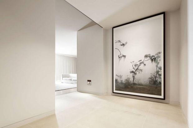 Minimal, bright interior hallway or entry area with large pale stone or porcelain floor tiles, smooth off-white walls, and a prominent oversized black-framed monochrome landscape artwork. An adjacent bright living or bedroom area is visible through the opening, with vertical wall detailing and light furnishings. The space has clean lines, sparse decoration, and a refined neutral palette.