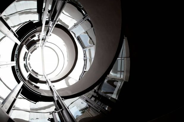 Upward-looking architectural view through a dramatic circular stair or atrium void. Curved concentric balconies, metal structural elements, reflective glazing, and a bright skylit opening create a strong spiral-like composition with high black-and-white contrast.