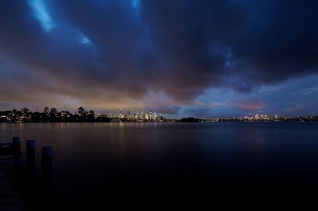A wide nighttime waterfront view across calm dark water toward a brightly illuminated city skyline. Dramatic blue and purple storm clouds fill the sky, with a dark silhouetted shoreline and a small dock or jetty structure in the lower-left foreground. No building interior or individual architectural subject dominates the image.