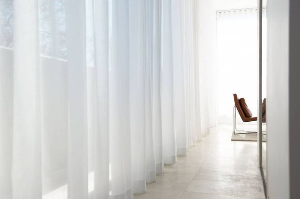Minimal, bright interior corridor or open-plan space lined with full-height sheer white curtains. Pale large-format flooring and extensive glazing create an airy, softly filtered atmosphere. A single modern brown leather chair with a slim metal frame is visible at the far end, with a partial reflection in the right-hand glazing.