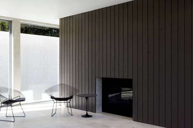 Minimalist contemporary interior with a dark vertical timber-panelled feature wall incorporating a built-in black fireplace. Bright clerestory glazing and white walls bring daylight into the space, with two wire lounge chairs and a small side table on a pale smooth floor.