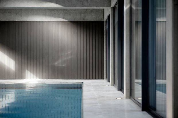 A refined indoor pool courtyard or pool area with a turquoise rectangular pool, pale stone or concrete paving, dark vertically ribbed wall panelling, and exposed concrete beams. Full-height glazed doors and windows line the right side, reflecting the pool and connecting to an adjacent interior. The composition is minimalist and architectural, with strong linear detailing and subdued natural light.