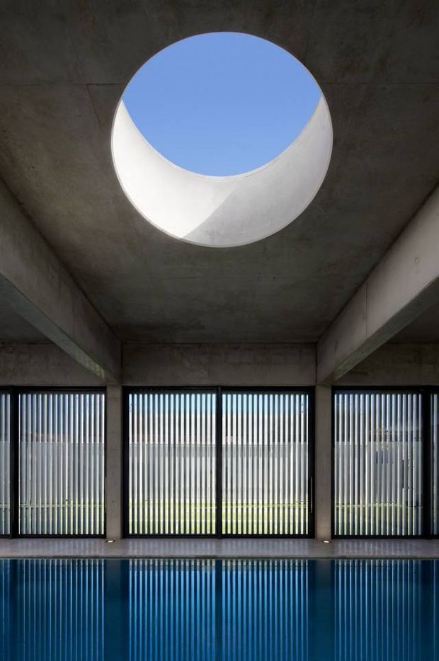 Interior view of a stark concrete pool area with a large circular skylight, exposed concrete beams and ceiling, and tall glazed openings fitted with vertical louvres. The blue water in the foreground reflects the louvred windows and daylight, creating a calm, graphic composition.
