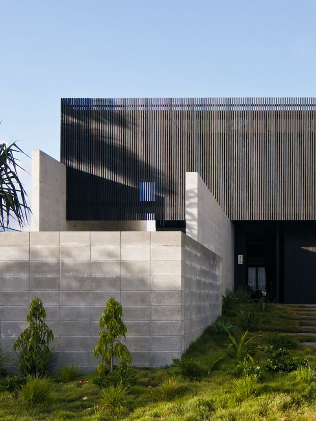 Exterior view of a contemporary, rectilinear building with a dark vertical slatted facade screening the upper volume. Pale rendered or concrete walls and a low blockwork boundary wall frame a recessed dark entry, with a landscaped garden and stepping path in the foreground.